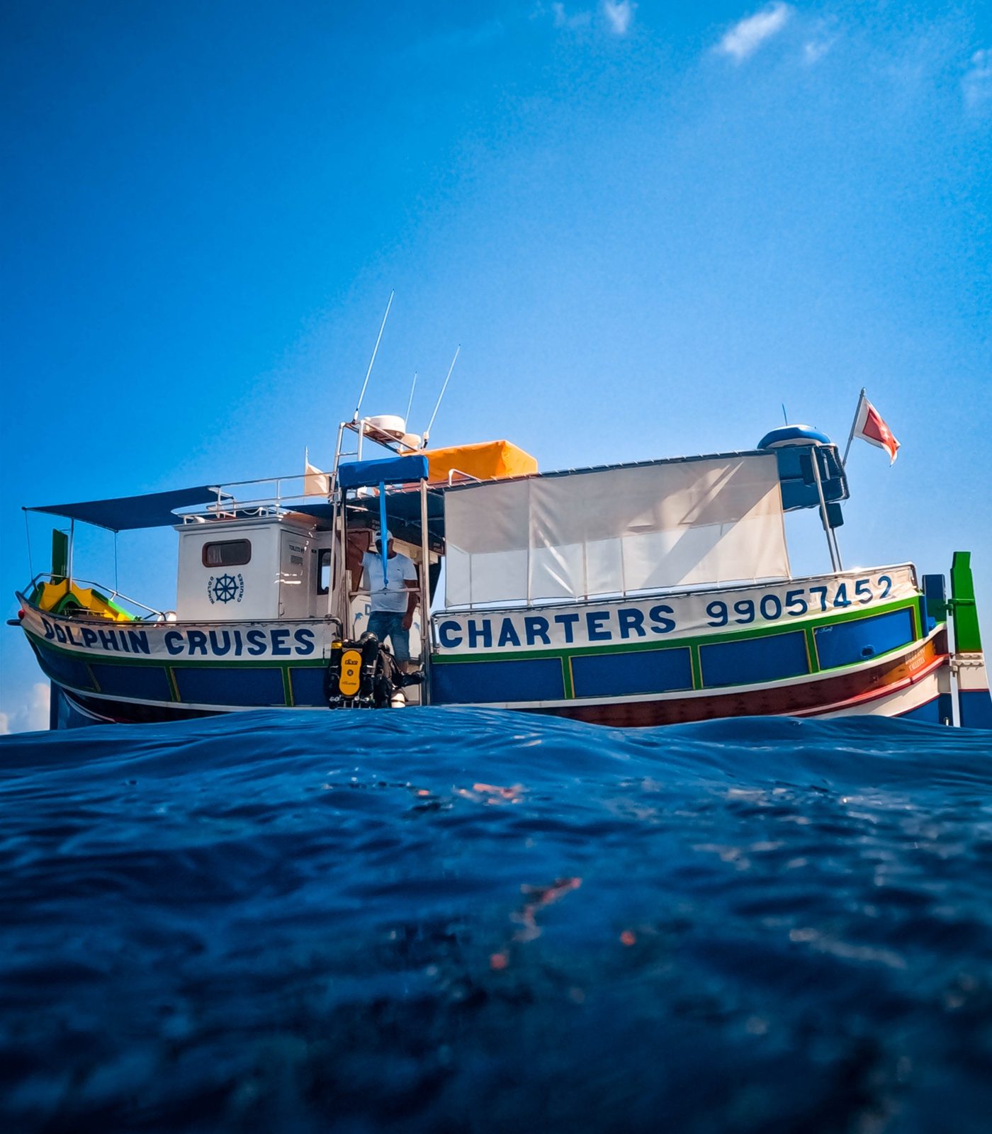 A traditional Maltese dive boat with a rebreather waiting at the stern and the captain on deck under a deep blue sky