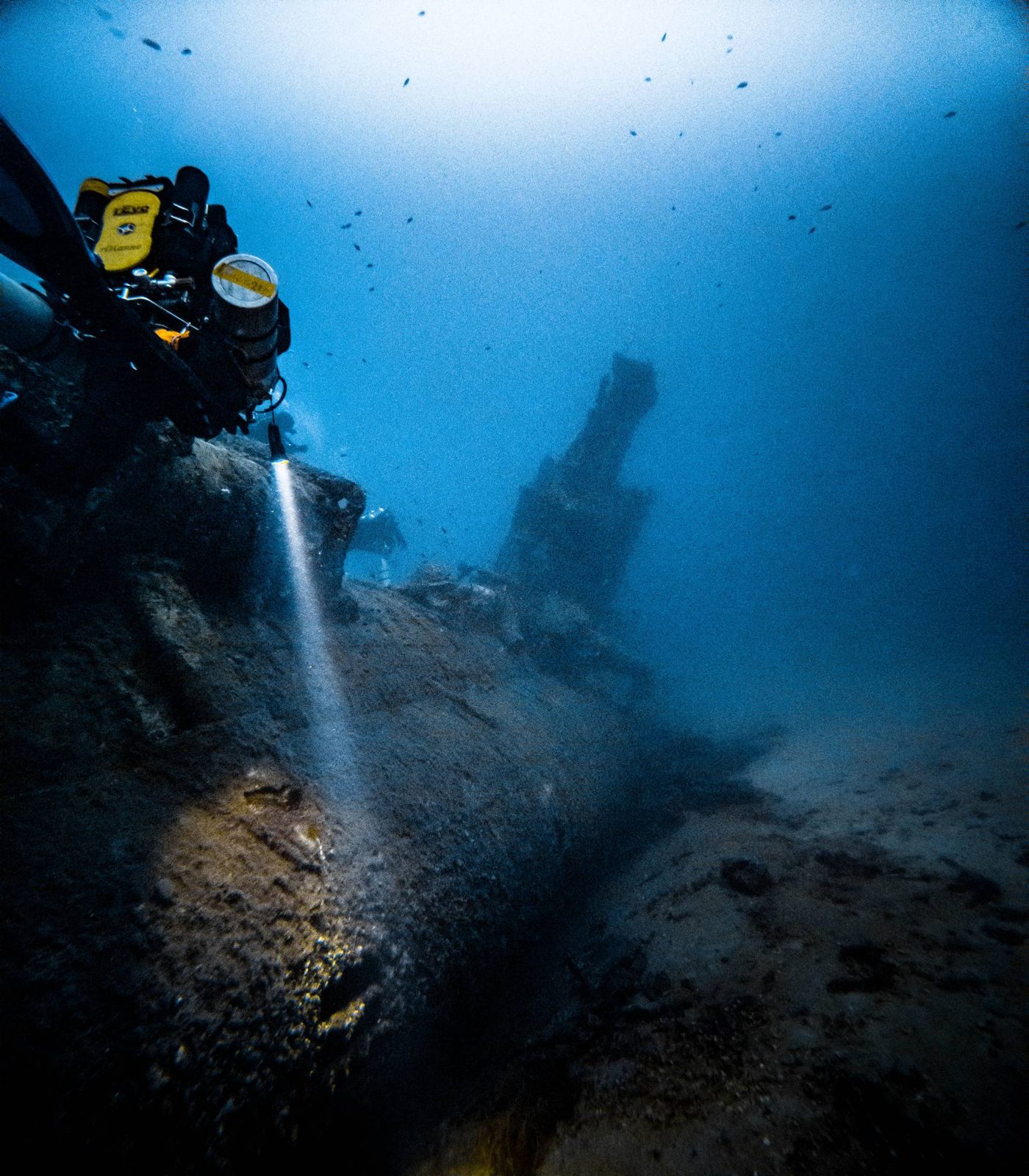 A rebreather diver lights the hull of a deep wreck with a torch, the rest of the wreck fading into the blue